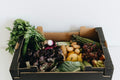Box of fresh vegetables including leafy greens, potatoes, and other produce on a white background